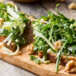 Close-up of apple tartines topped with arugula, walnuts, and julienned apples on toasted bread, served on a wooden board.