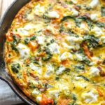 Butternut squash frittata in skillet on wooden table - Lightly browned edges with visible kale, bacon, and creamy goat cheese throughout.