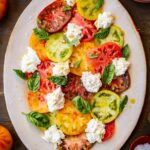 Overhead shot of completed Caprese salad on a platter, surrounded by fresh tomatoes, basil, and a bowl of flaky salt.