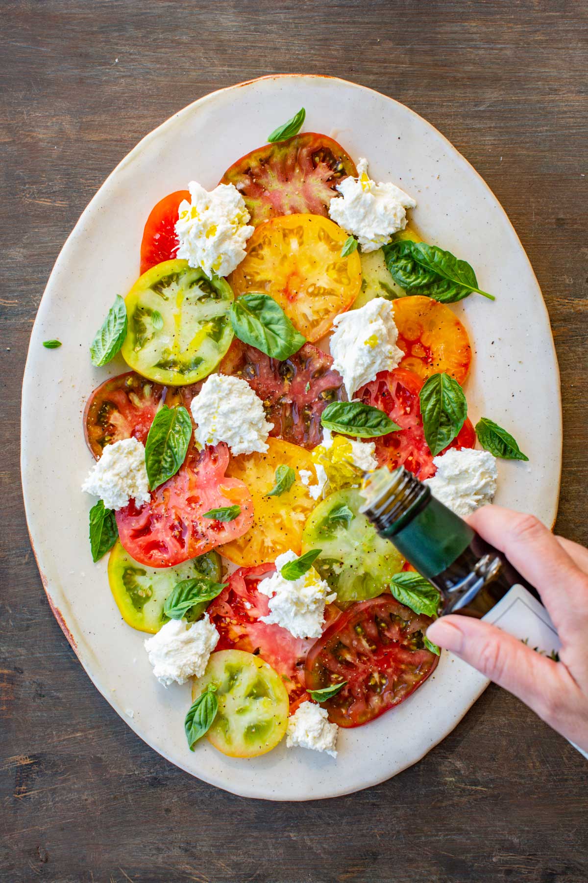 Caprese salad being assembled with basil leaves scattered over tomatoes and mozzarella, while olive oil is drizzled on top.