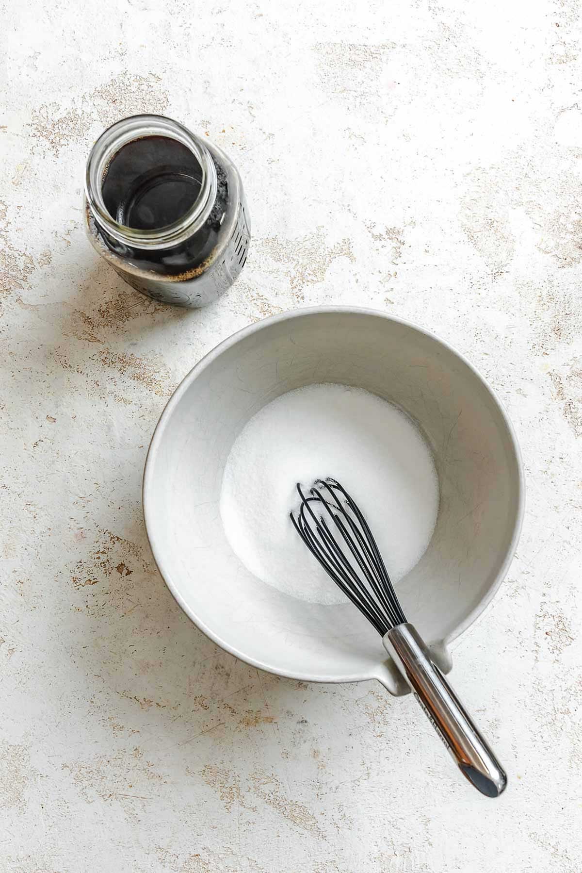 Bowl of sugar with whisk next to a jar of freshly brewed coffee on a light background.