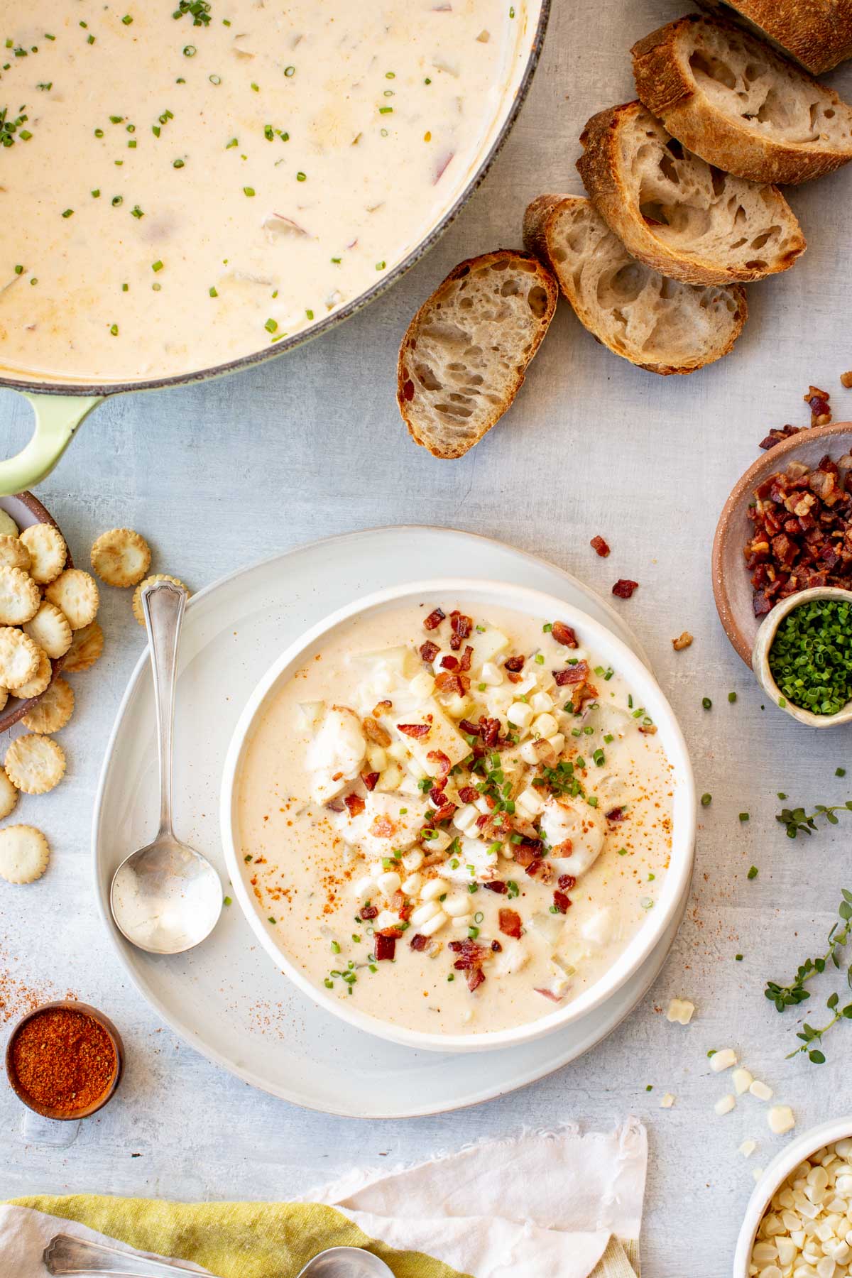 Overhead shot of a pot of corn and crab chowder sprinkled with chives, surrounded by sliced crusty bread, a bowl of oyster crackers, bacon crumbles, and fresh chives, with a plated bowl of chowder garnished and ready to serve.