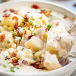 Close-up of a spoonful of creamy chowder lifted from a bowl, showing chunks of potato, sweet corn, bacon, and crab meat in a rich broth with bread in the background.
