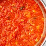 Close-up of fresh tomato sauce simmering in a stainless steel pan, with garlic slices and basil visible in the sauce.