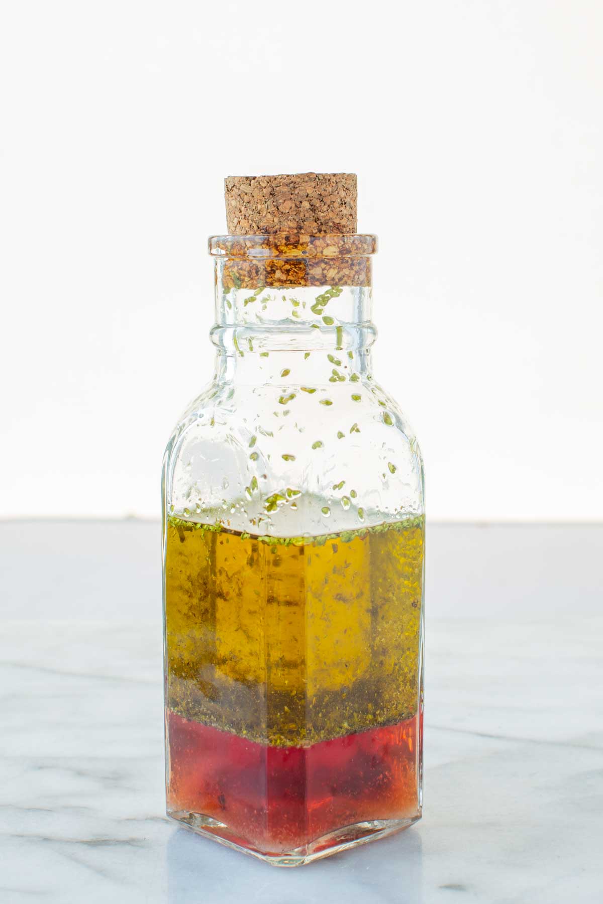 A clear glass bottle with a cork top filled with homemade Italian dressing, showing layers of red wine vinegar and extra virgin olive oil with dried herbs floating on top.