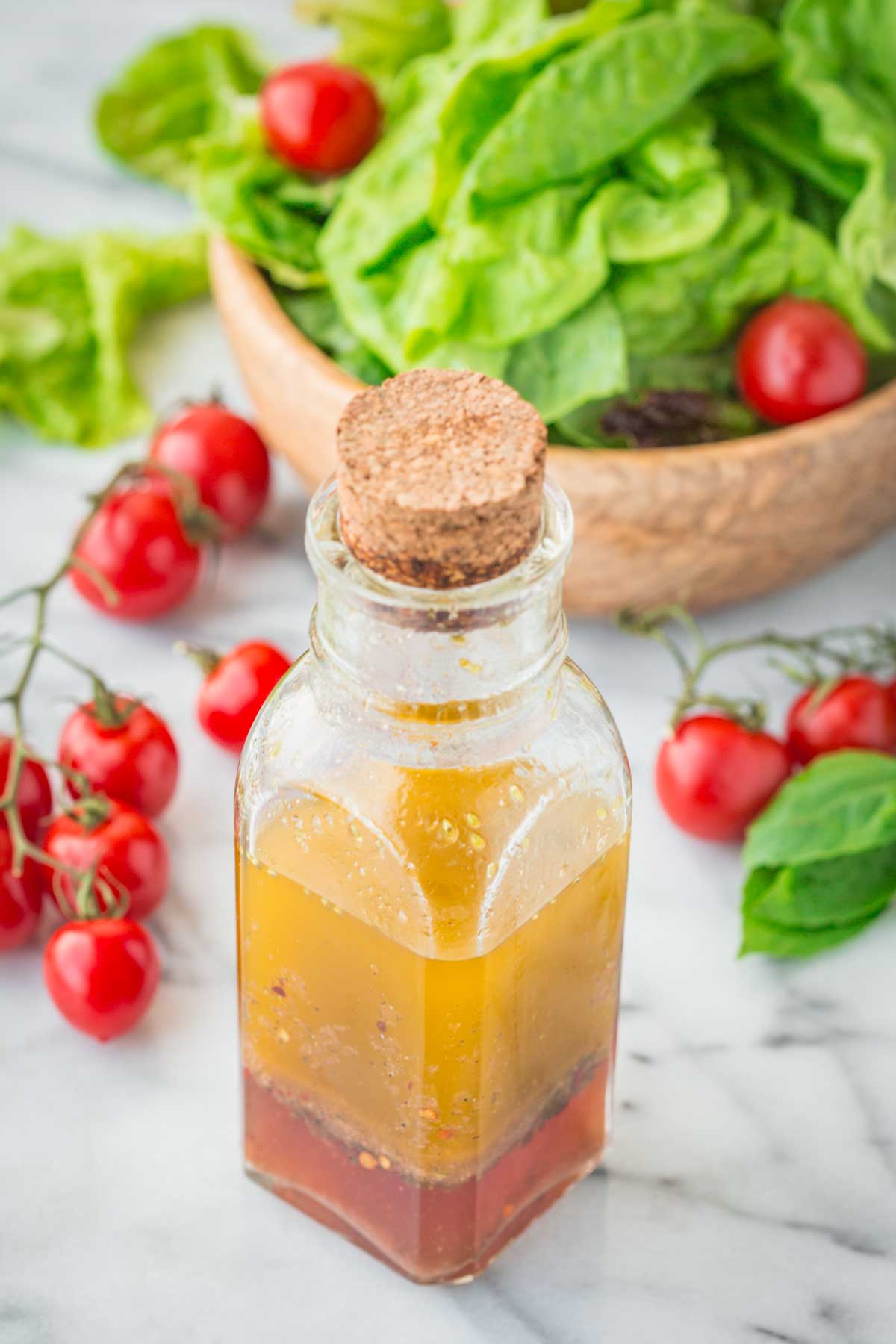 Homemade Italian dressing in a corked glass bottle placed on a marble surface with a wooden bowl of lettuce, fresh basil, and vine-ripened cherry tomatoes in the background.