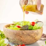 Hand pouring homemade Italian vinaigrette from a glass bottle over a wooden bowl filled with green salad and cherry tomatoes.