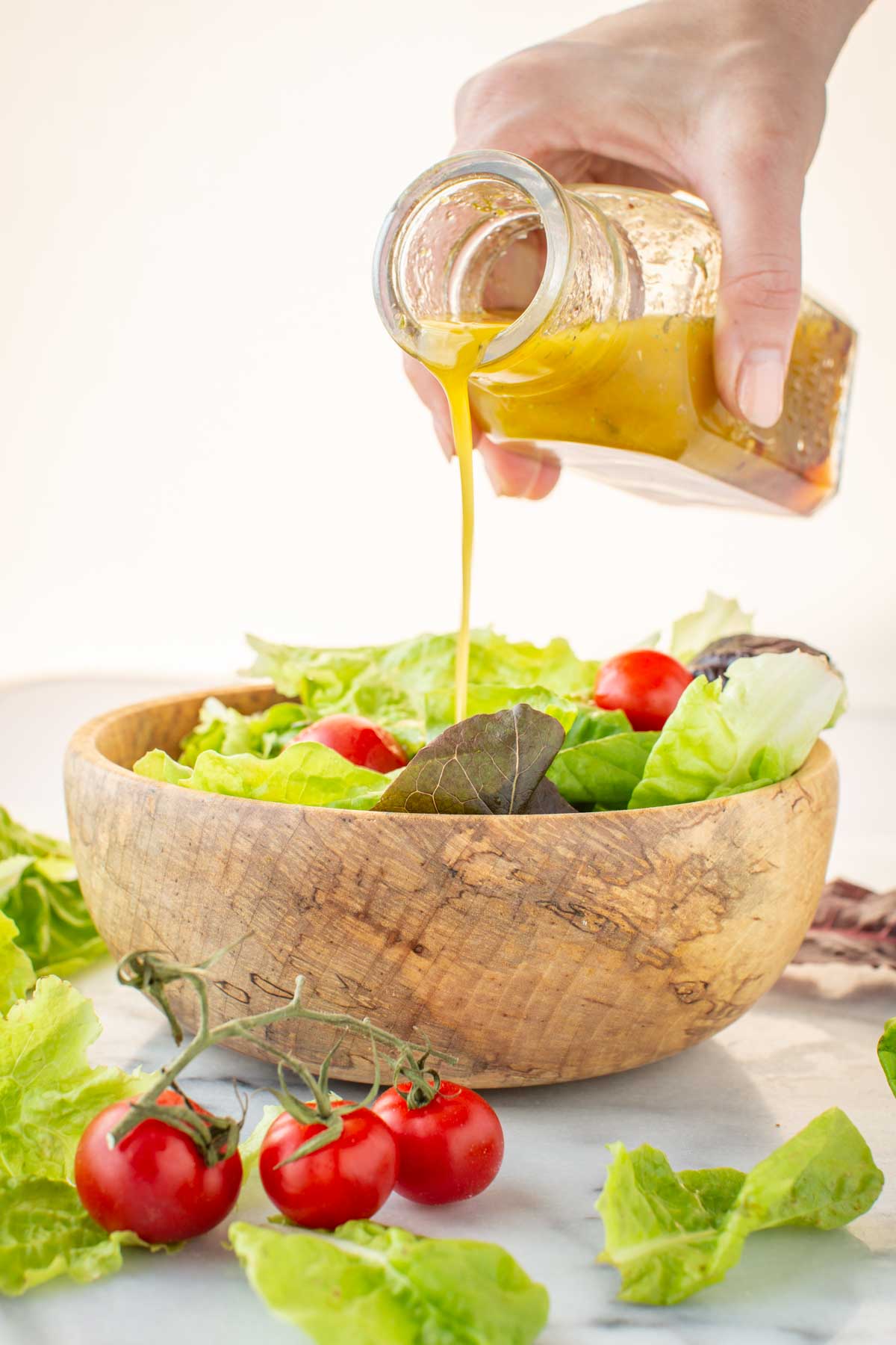 Hand pouring homemade Italian vinaigrette from a glass bottle over a wooden bowl filled with green salad and cherry tomatoes.