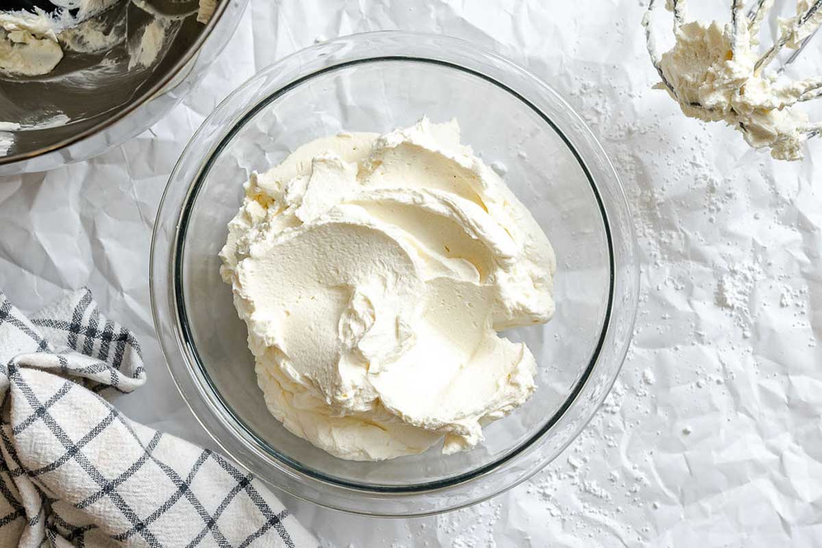 Large glass bowl filled with freshly whipped mascarpone cream on a textured white background, with stand mixer and whisk attachment nearby.