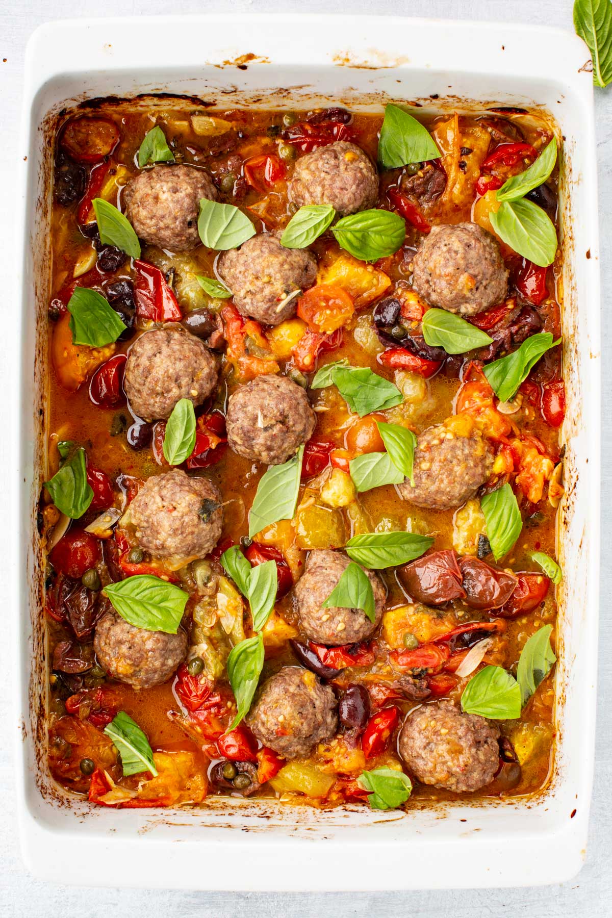 White baking dish filled with roasted meatballs in a chunky tomato puttanesca sauce, garnished with fresh basil leaves.