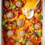 Partially eaten casserole dish of roasted meatballs with tomato sauce, olives, and basil, with a spoon resting inside.