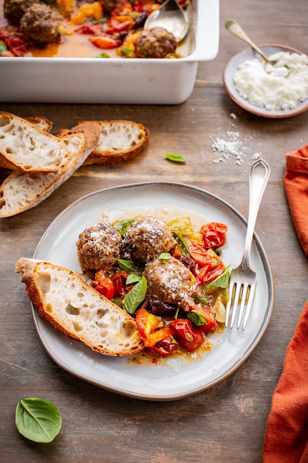 Meatballs with puttanesca sauce plated with bread, fresh basil, and grated cheese, with the casserole dish in the background.