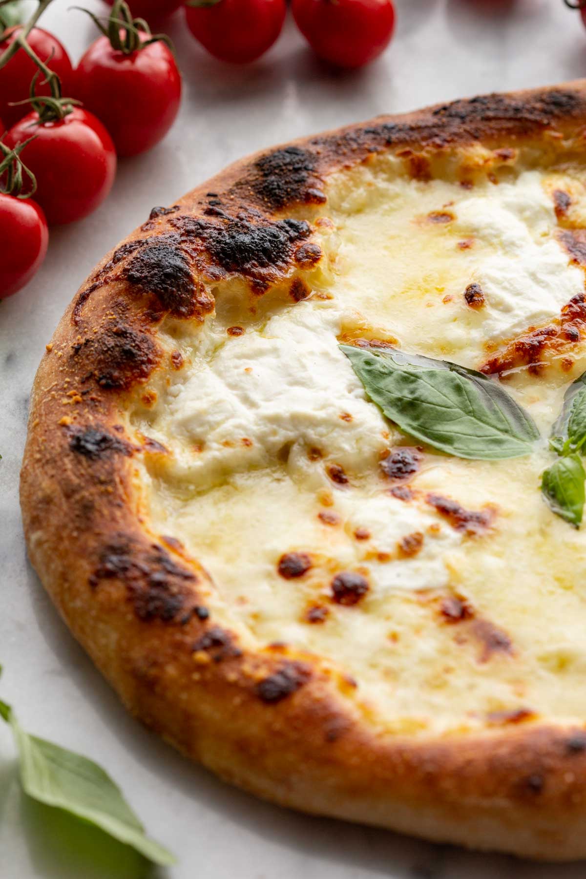 Side angle pizza - White pizza with blistered crust and melted cheeses, styled with a bunch of cherry tomatoes in the background.