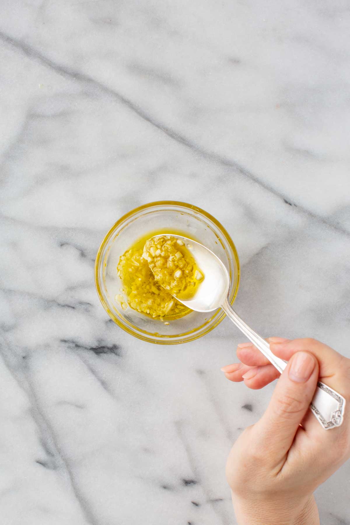 Garlic oil prep - A hand holding a spoon mixing olive oil and minced garlic in a small glass bowl on a marble counter.