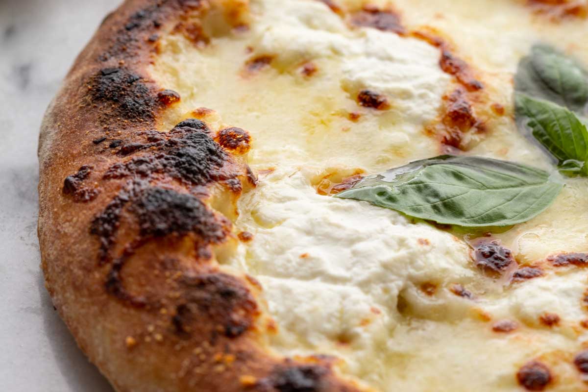 Close-up cheese detail – Close view of bubbling ricotta and melted mozzarella with charred crust and a basil leaf on top.