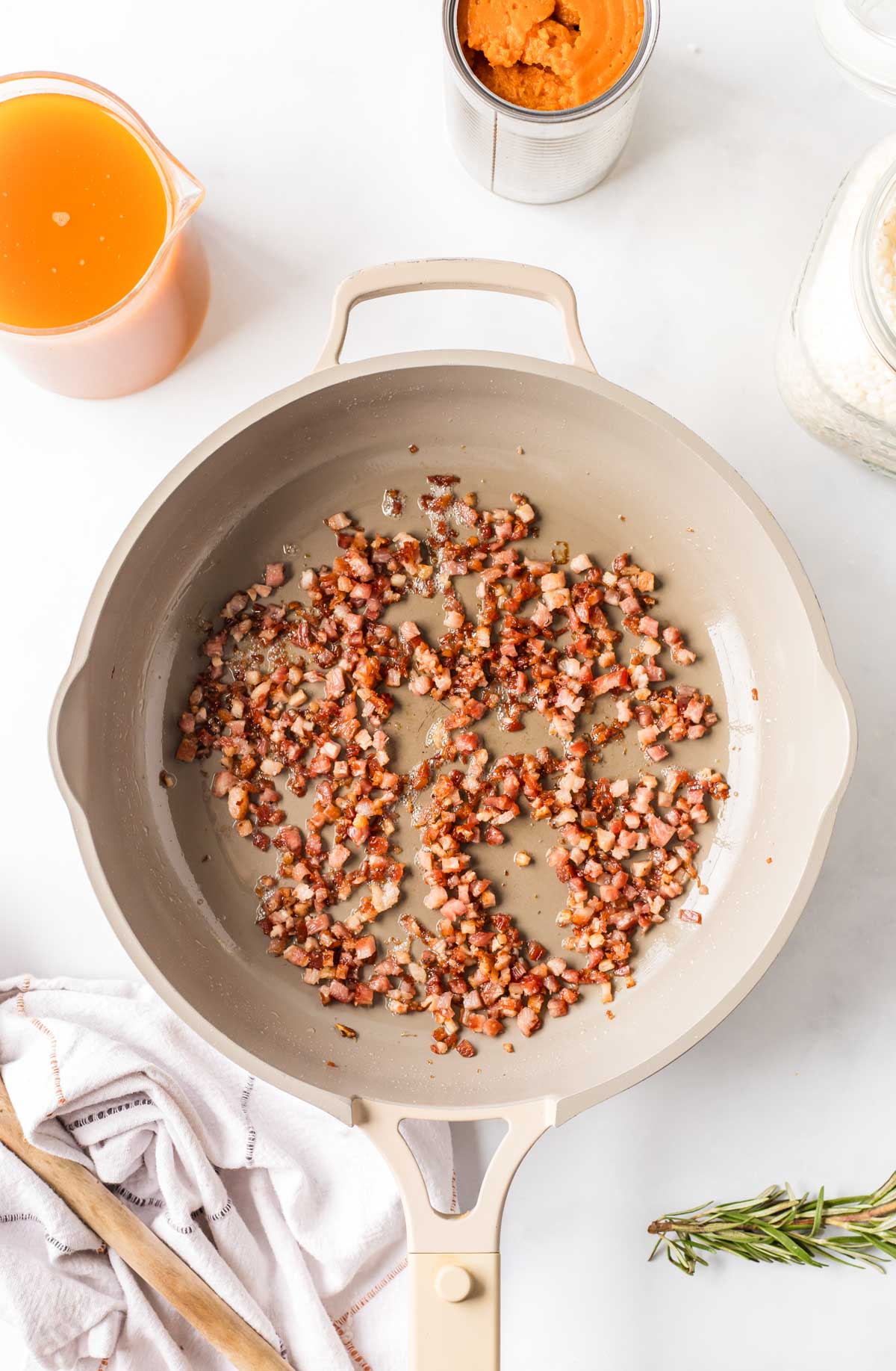 Crisped pancetta in a beige skillet, with a can of pumpkin puree, a jar of arborio rice, and a measuring cup of vegetable stock on the counter.