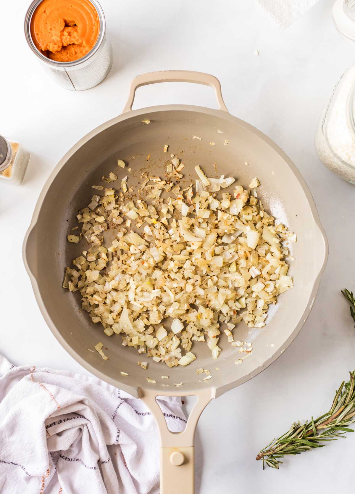Golden sautéed onions and garlic in a skillet, with a can of pumpkin and a sprig of fresh rosemary nearby.