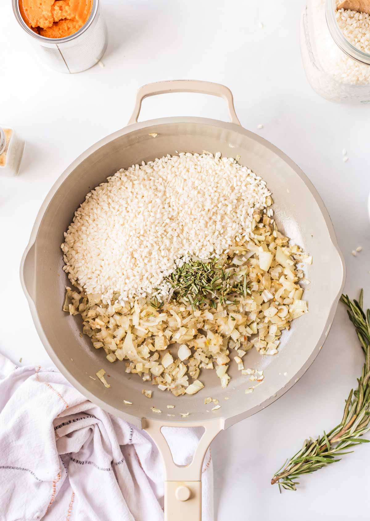 Uncooked arborio rice and chopped rosemary added to sautéed onions in a skillet, ready to toast.