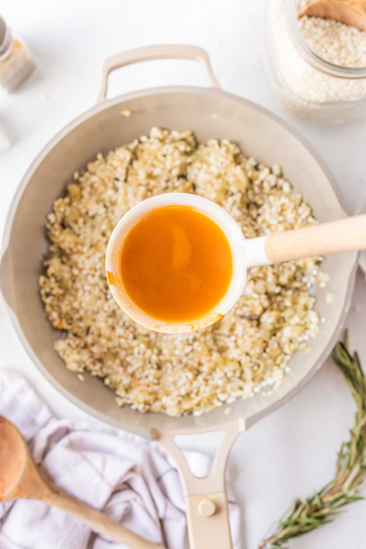A ladle of warm vegetable stock held over a skillet of toasted rice, about to be stirred in.