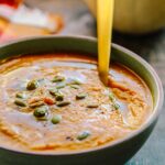 A close-up bowl of creamy pumpkin soup topped with toasted pumpkin seeds and a drizzle of olive oil, with a gold spoon resting inside.