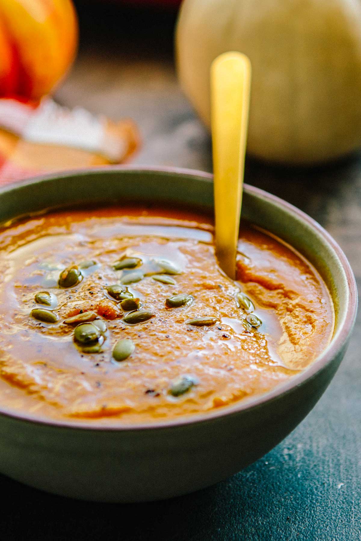 A close-up bowl of creamy pumpkin soup topped with toasted pumpkin seeds and a drizzle of olive oil, with a gold spoon resting inside.