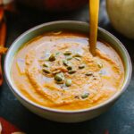 Wide shot of pumpkin soup in a bowl with a spoon, sprinkled with seeds and olive oil, with pumpkins and a fall napkin in the background.