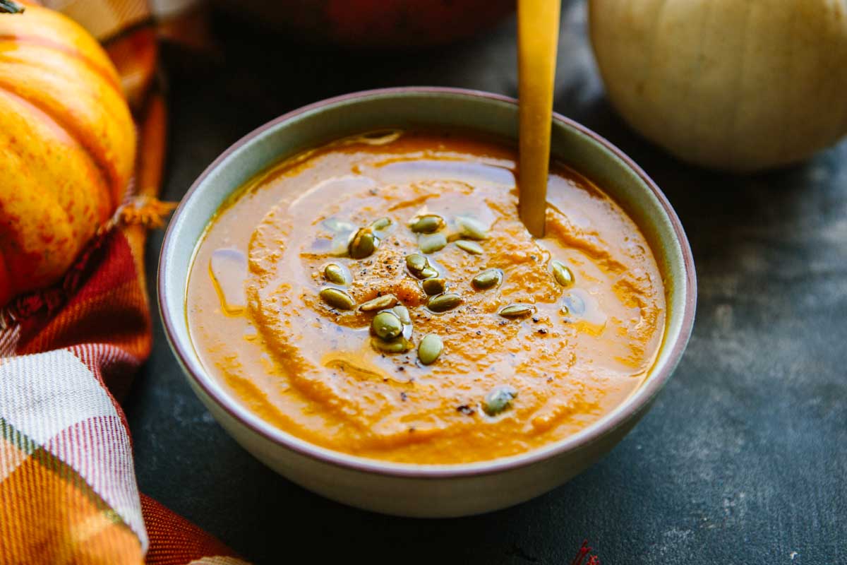 Wide shot of pumpkin soup in a bowl with a spoon, sprinkled with seeds and olive oil, with pumpkins and a fall napkin in the background.