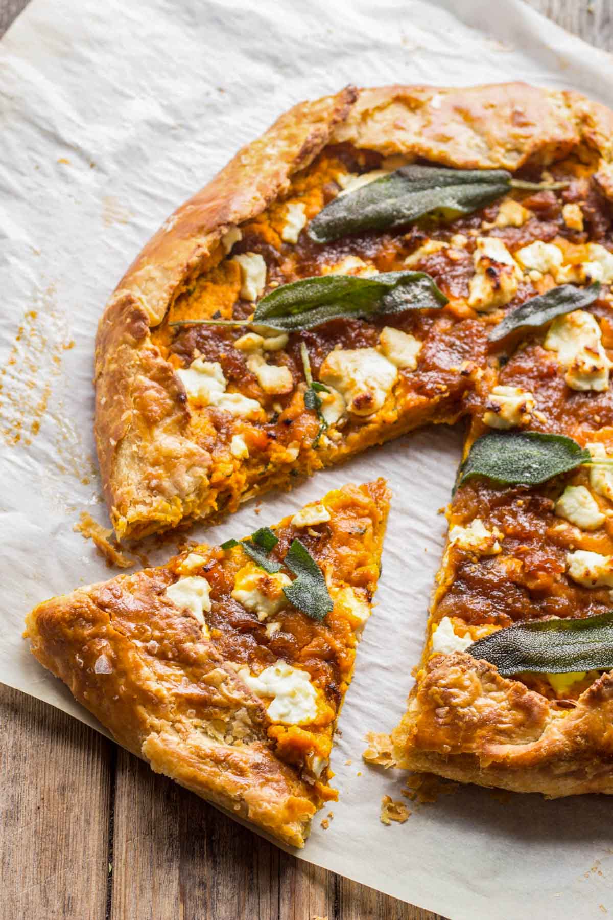 Overhead view of a pumpkin and goat cheese galette on parchment paper, showing its flaky edges, browned onions, creamy cheese, and sage leaves with a slice pulled away.