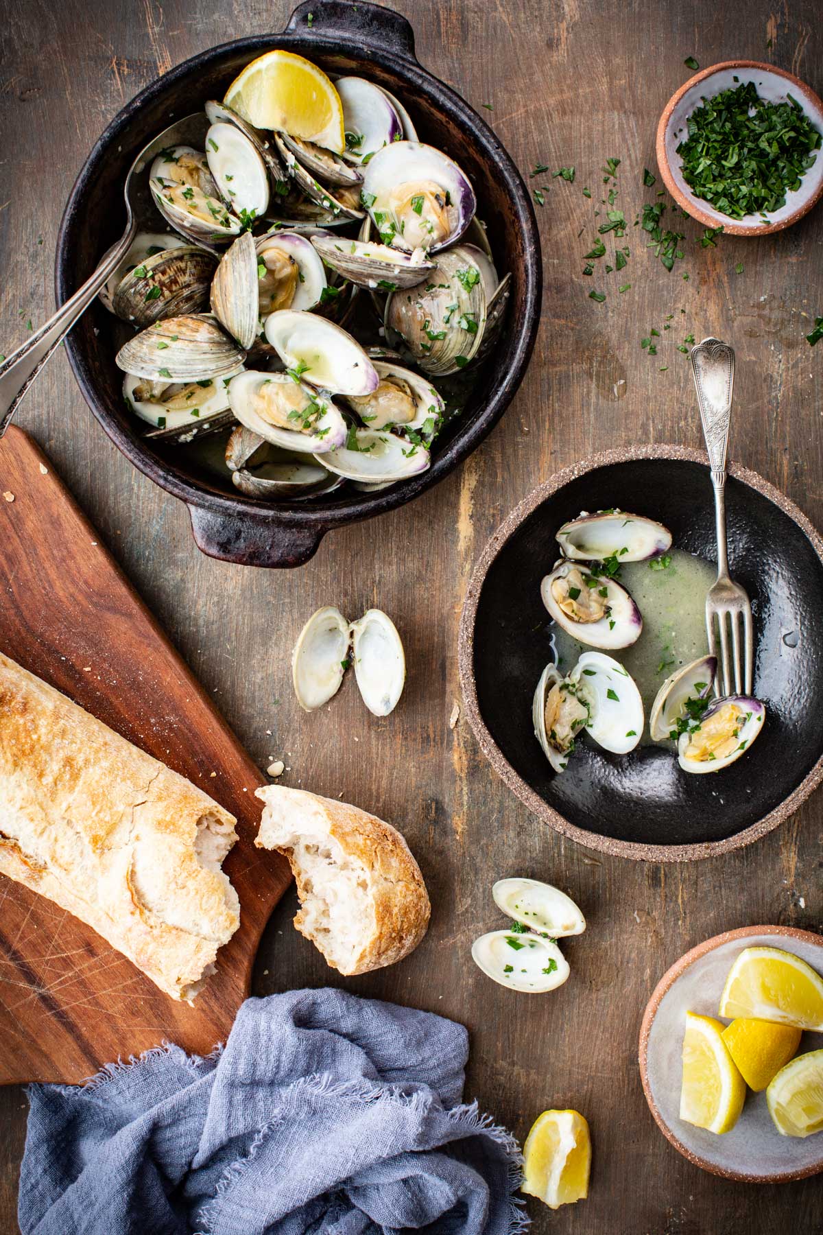 Finished clams with bread overhead - A rustic black bowl filled with steamed clams in garlic butter broth, garnished with parsley and lemon wedges, set on a wooden table with torn crusty bread and scattered clam shells.