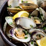 A detailed view of opened clams in their shells, sprinkled with parsley and served with lemon wedges in a rustic black bowl.