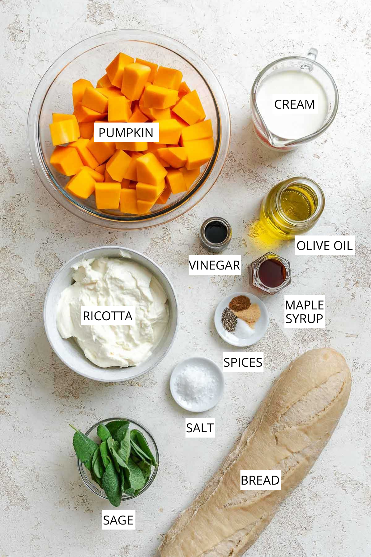Overhead view of labeled pumpkin ricotta crostini ingredients including cubed pumpkin, ricotta, cream, olive oil, balsamic vinegar, maple syrup, sage, salt, bread, and spices.