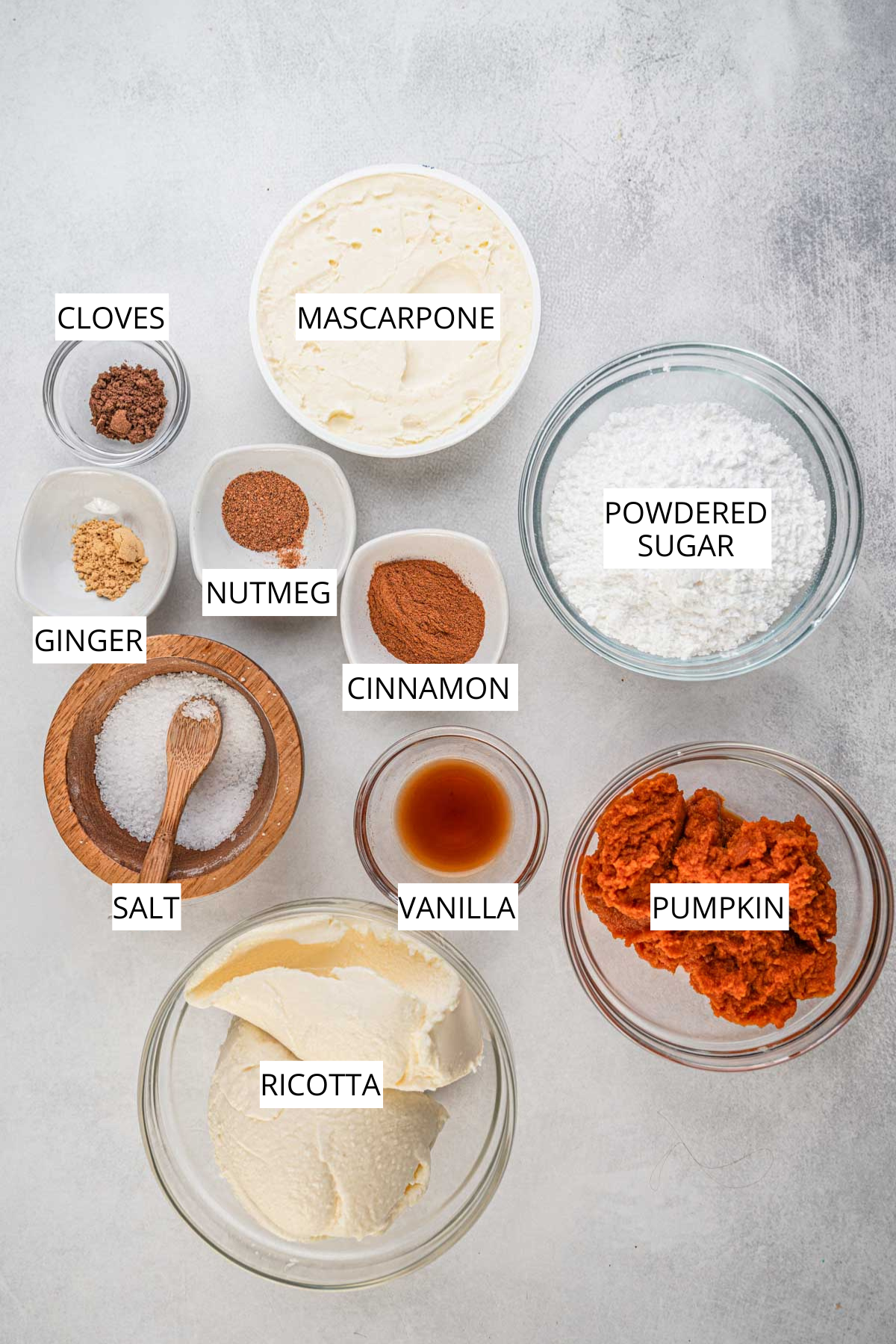 Overhead image showing labeled ingredients for pumpkin cannoli filling including ricotta, mascarpone, pumpkin puree, powdered sugar, vanilla, salt, cinnamon, nutmeg, ginger, and cloves.