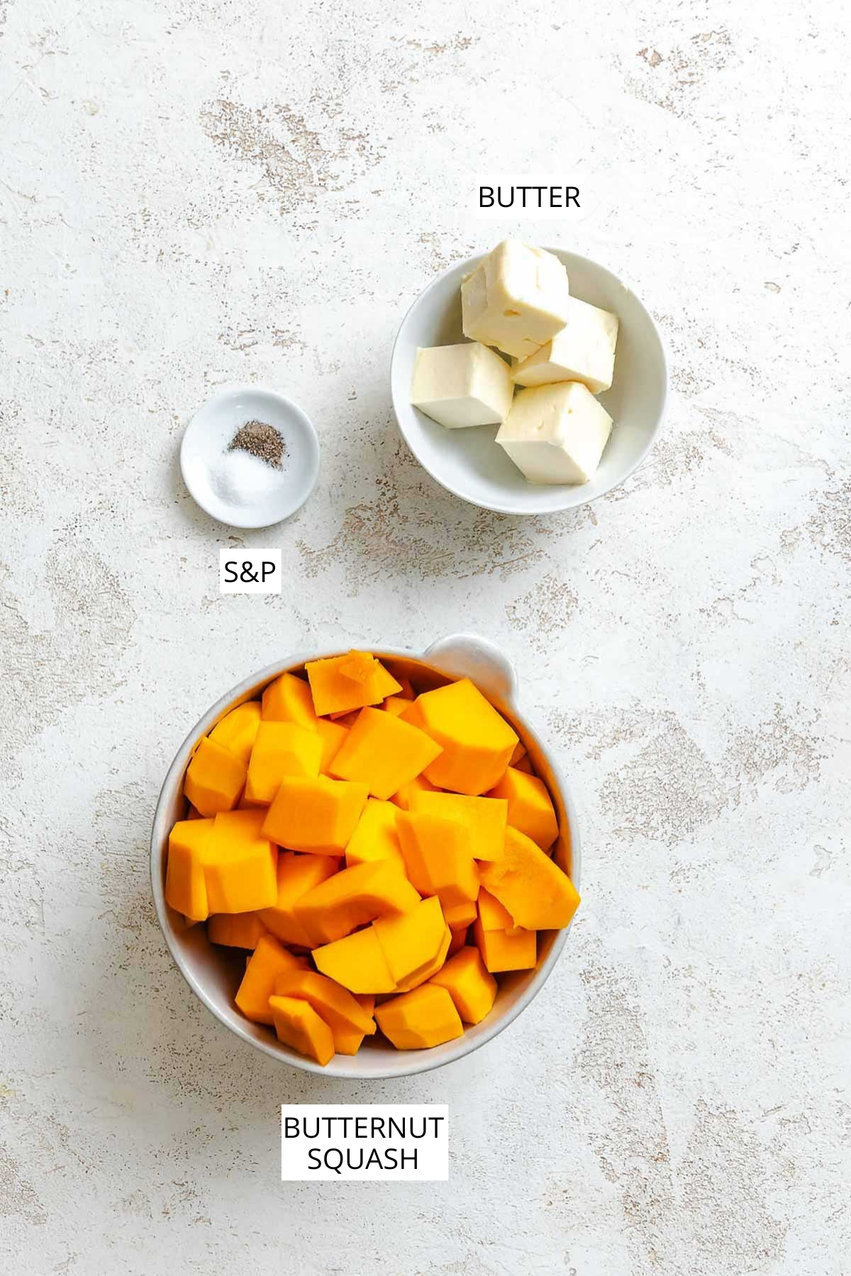 Overhead view of labeled ingredients for brown butter squash puree, including cubed butternut squash, butter, salt, and pepper on a light surface.