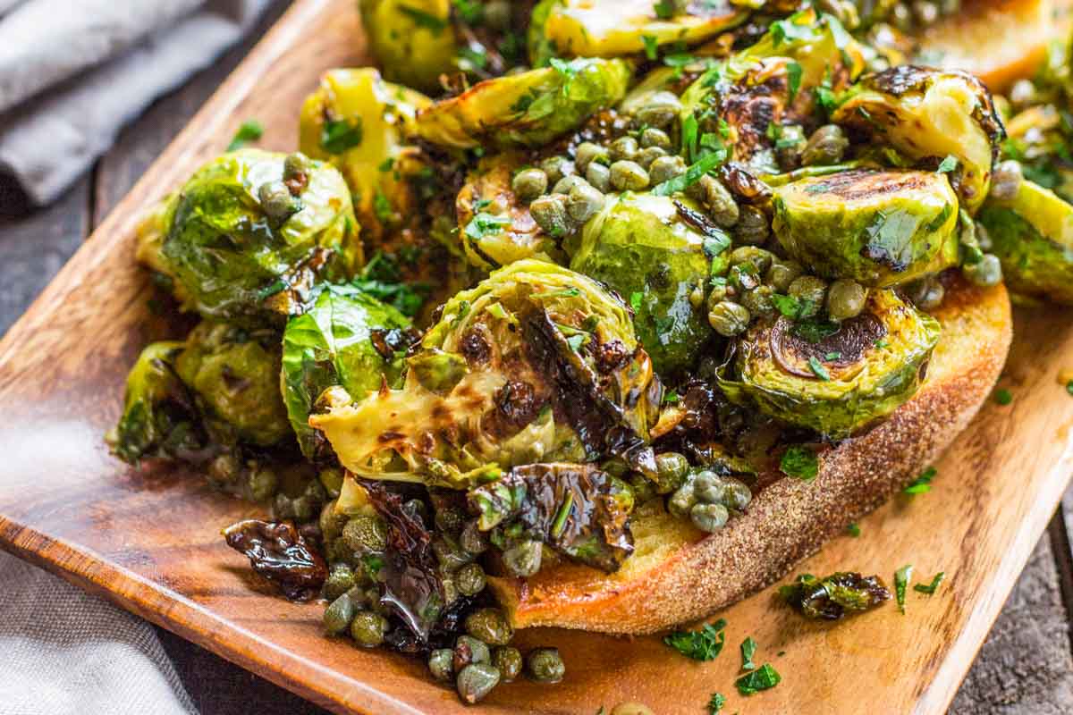 Close-up of crispy Brussels sprouts and briny capers on toasted bread, coated in glossy lemon-anchovy butter sauce.
