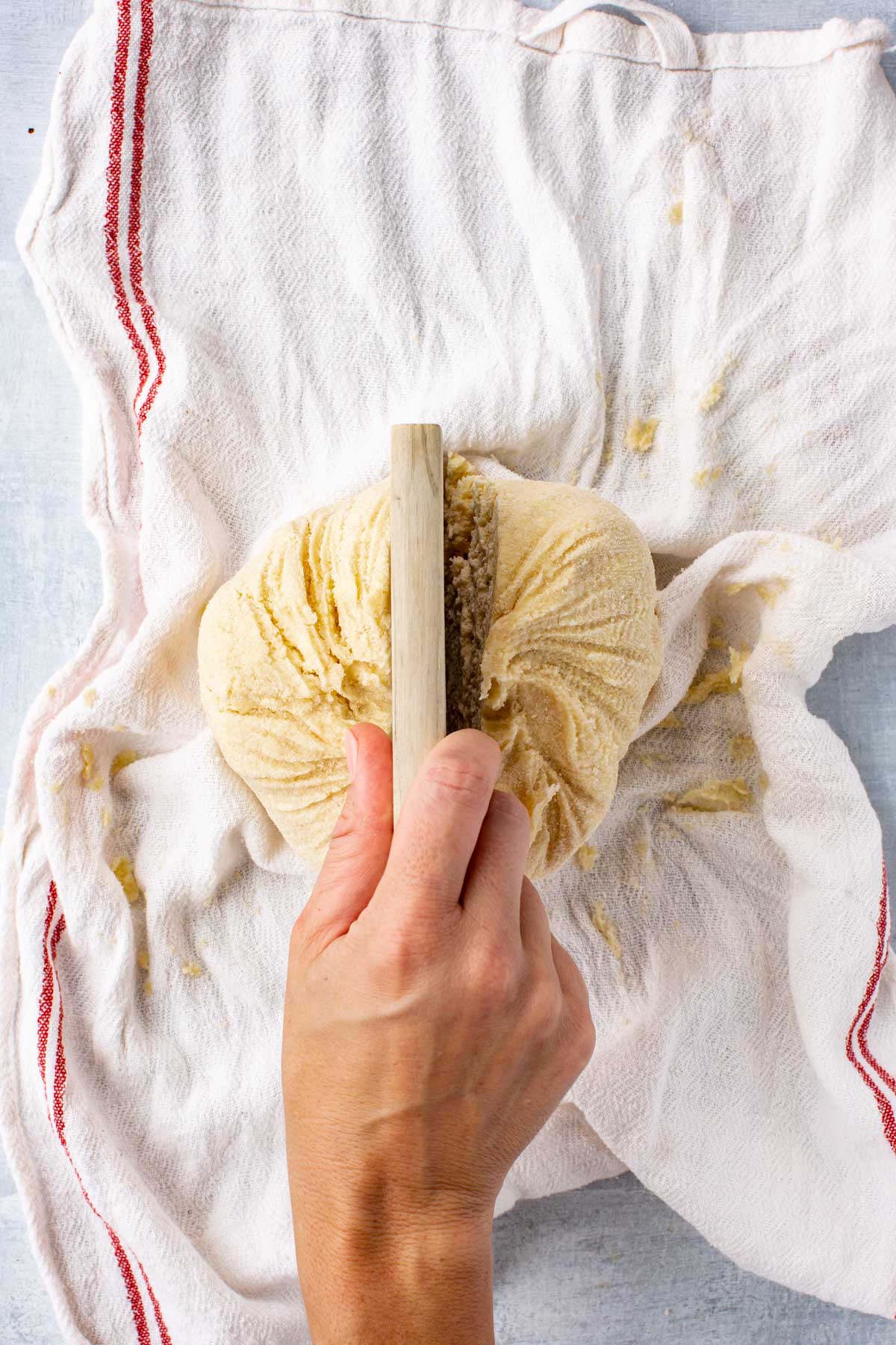 A hand uses a bench scraper to divide freshly formed buttermilk pie dough wrapped tightly in a white tea towel.