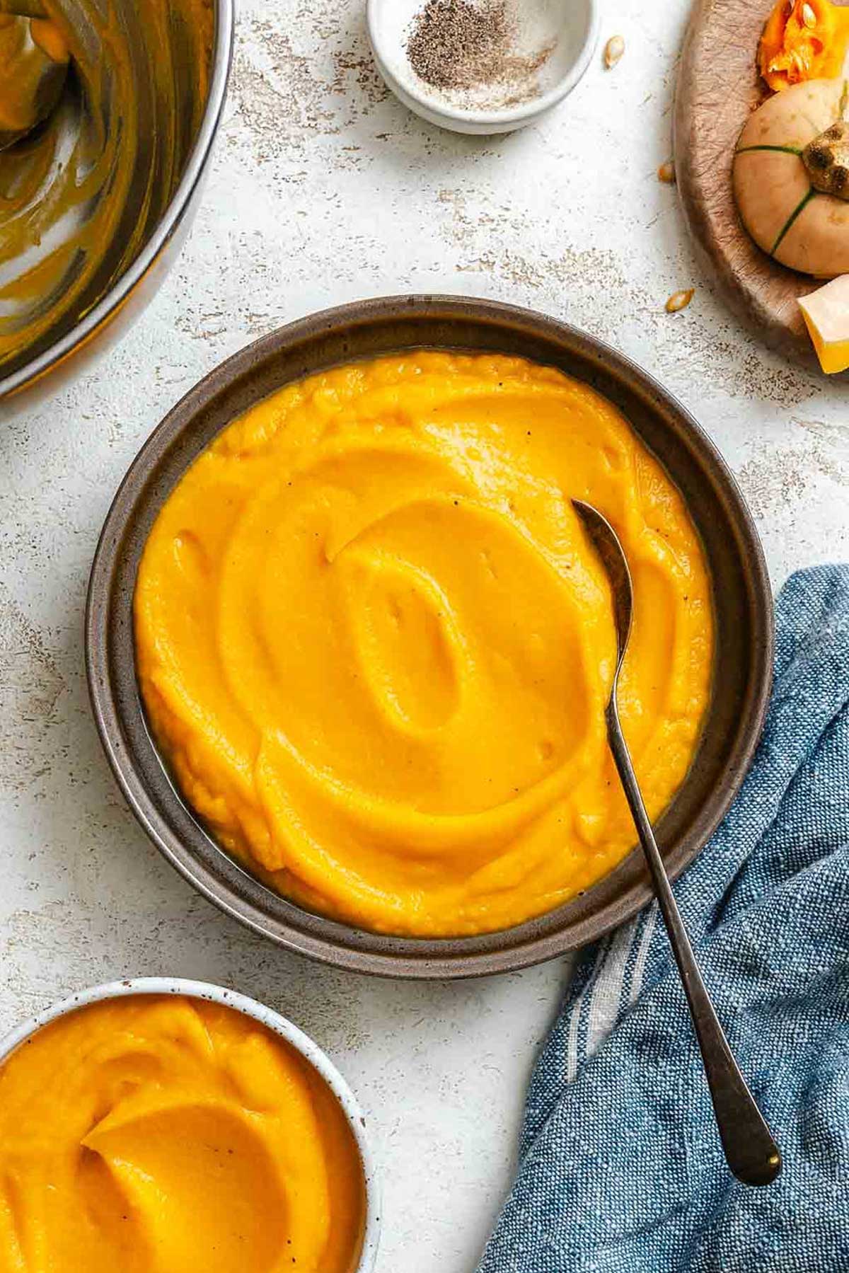 Bowl of bright orange brown butter squash puree with a spoon, surrounded by bowls of seasoning and a blue linen napkin.