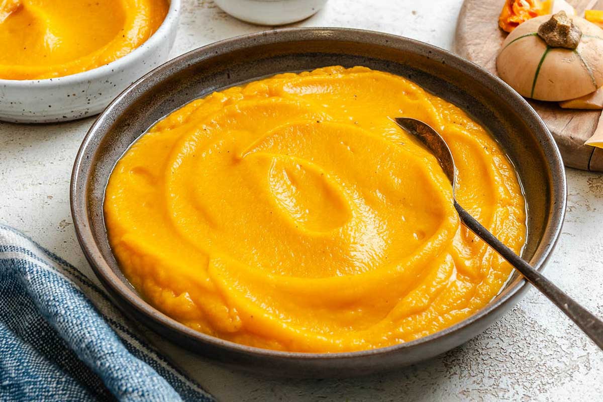 Wide shot of two bowls filled with brown butter squash puree, one large and one small, placed on a textured white surface with a cloth napkin.
