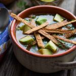 A warm ceramic bowl of chicken tortilla soup topped with avocado chunks and crunchy tortilla strips, set next to a spoon on a colorful woven napkin.