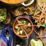 A rustic bowl of chicken tortilla soup topped with crispy tortilla strips and chunks of avocado, surrounded by lime wedges, extra strips, and a Dutch oven full of soup.