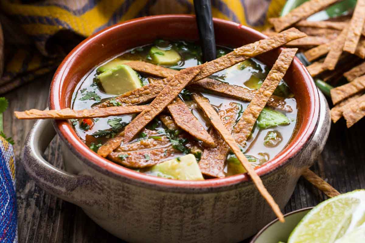 Close-up of chicken tortilla soup in a clay bowl, garnished with golden tortilla strips, avocado cubes, and fresh cilantro in a flavorful broth.