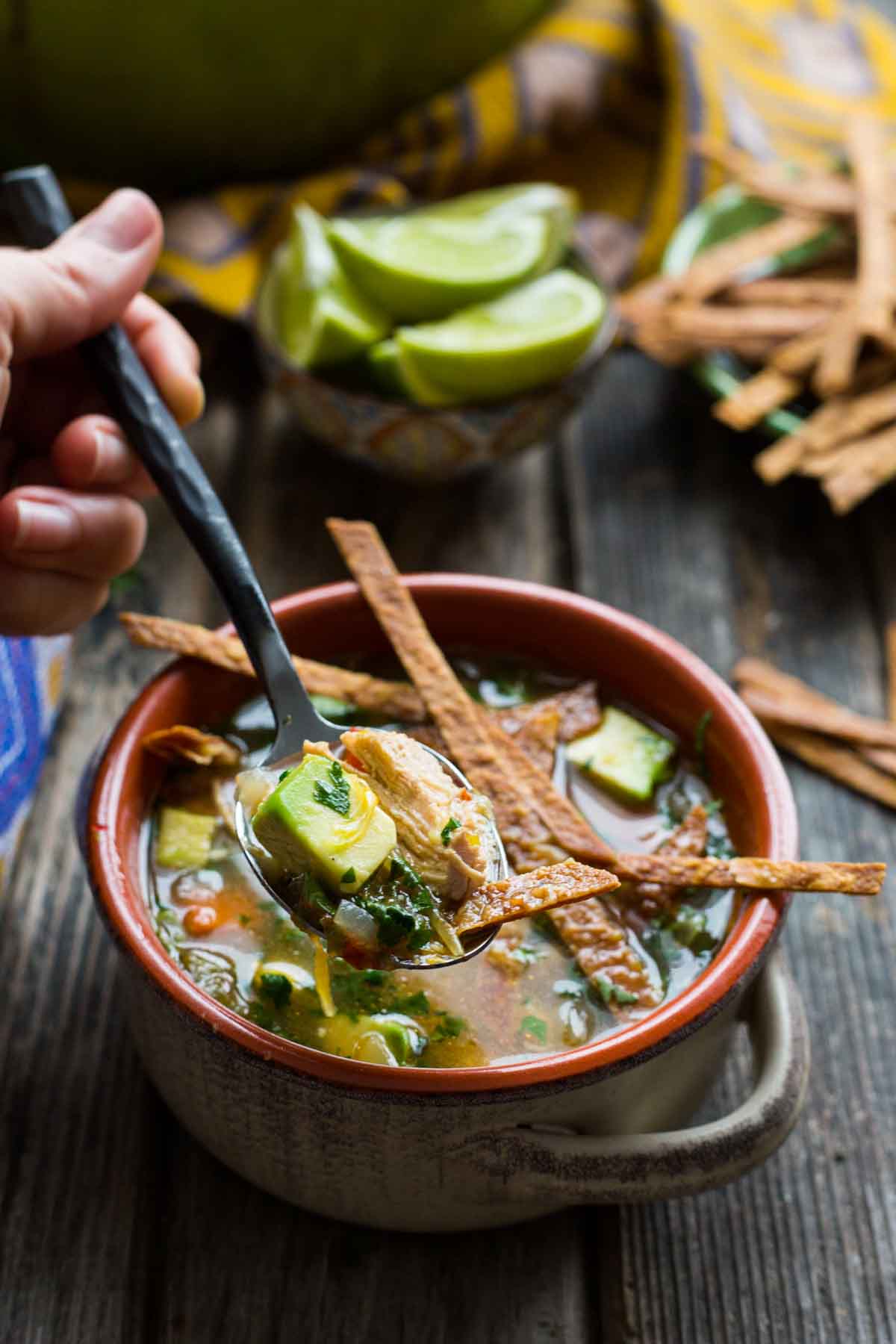 A hand holds a spoonful of chicken tortilla soup, lifting shredded chicken, avocado, and cilantro from a bowl topped with tortilla strips.