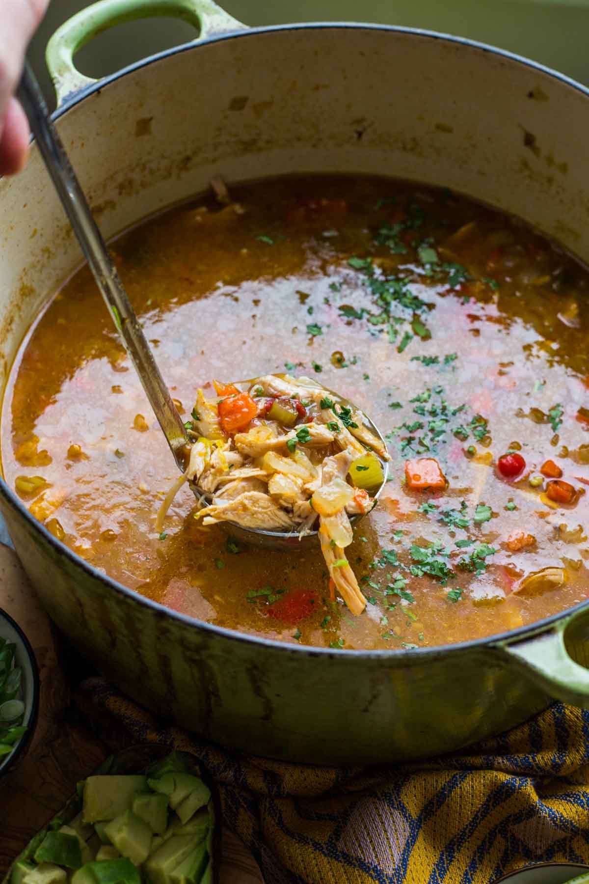 A ladle filled with shredded chicken, corn, and vegetables being lifted from a green Dutch oven full of steaming tortilla soup.