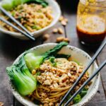 Two bowls of Dan Dan noodles with chili oil jar and bok choy on a rustic wooden table.
