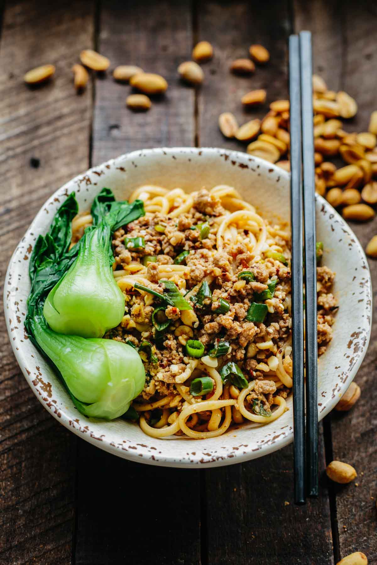 Bowl of Dan Dan noodles with chopsticks resting on the rim, surrounded by peanuts and bok choy.