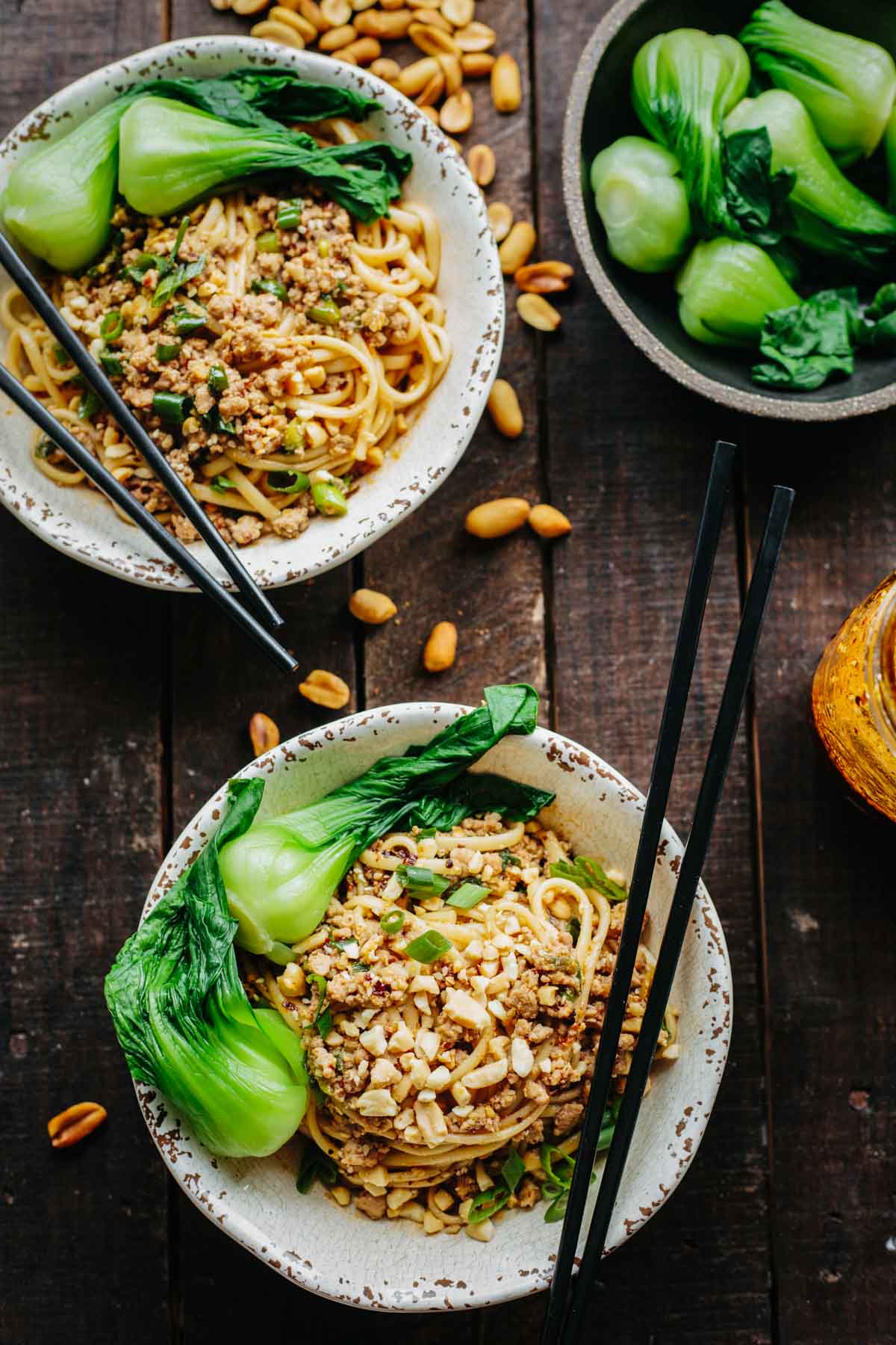 Overhead shot of two bowls of Dan Dan noodles with peanuts scattered around and a bowl of steamed bok choy on the side.
