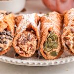 Four pumpkin cannoli on a white plate, each filled with creamy pumpkin ricotta filling and topped with chocolate chips, nuts, and pumpkin seeds.