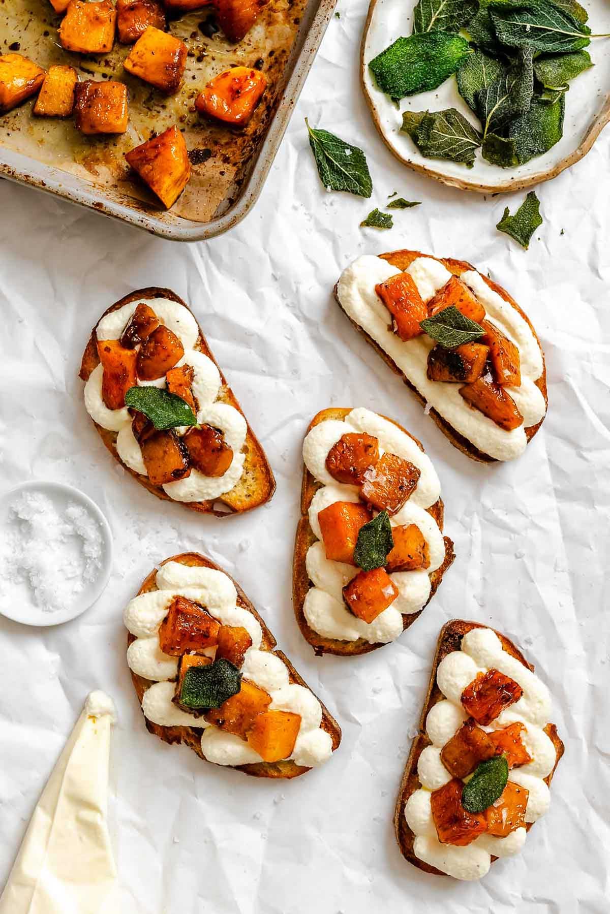 Assembling crostini with piped whipped ricotta, roasted pumpkin cubes, and crispy sage leaves on a sheet of parchment paper.