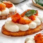 Close-up of a single pumpkin ricotta crostini showing creamy ricotta topping, caramelized pumpkin pieces, and a crisp sage leaf garnish.