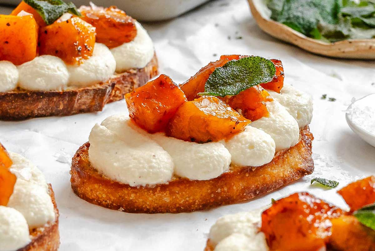 Close-up of a single pumpkin ricotta crostini showing creamy ricotta topping, caramelized pumpkin pieces, and a crisp sage leaf garnish.