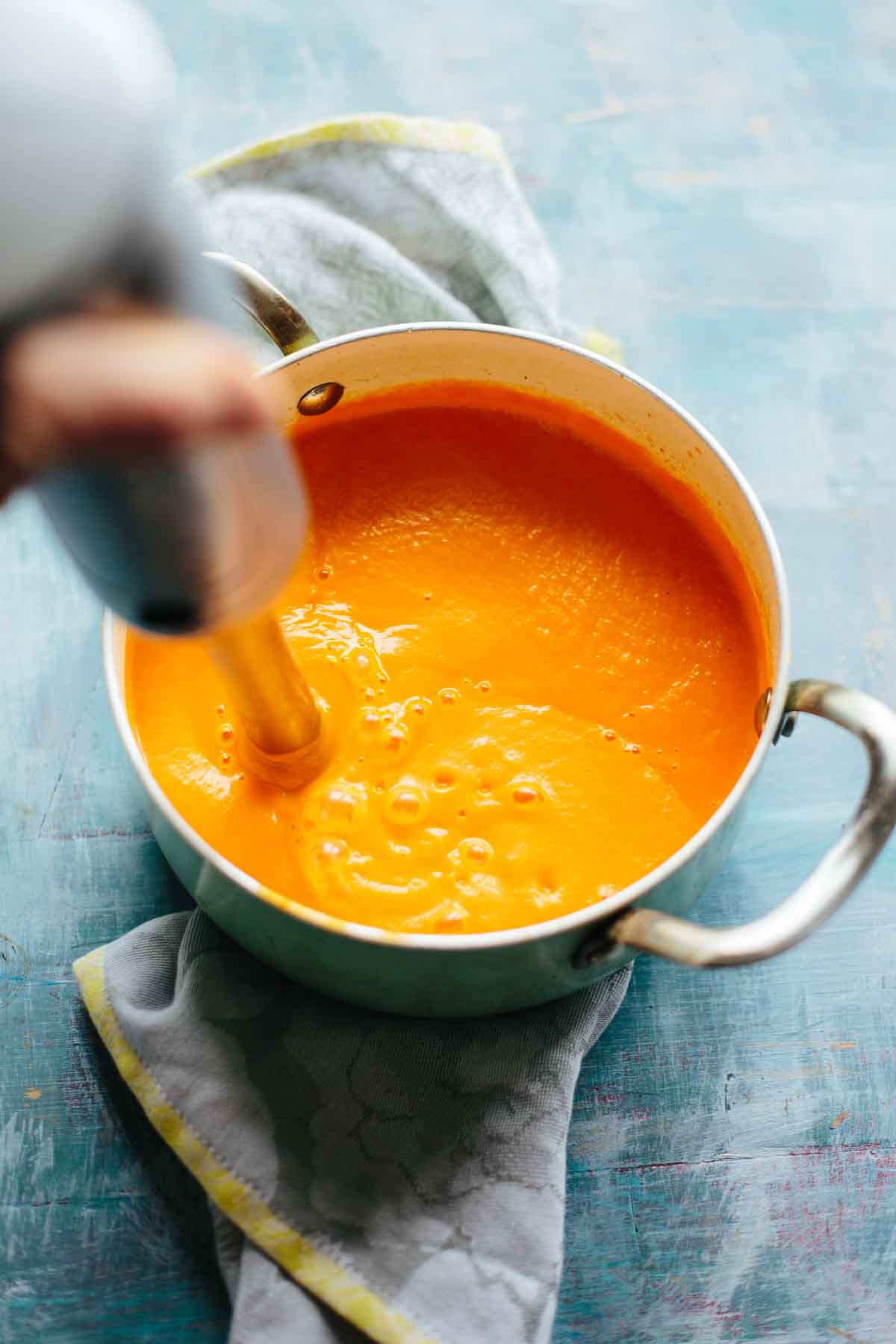 Bright orange spicy carrot soup being blended smooth with an immersion blender in a large pot on a blue surface.